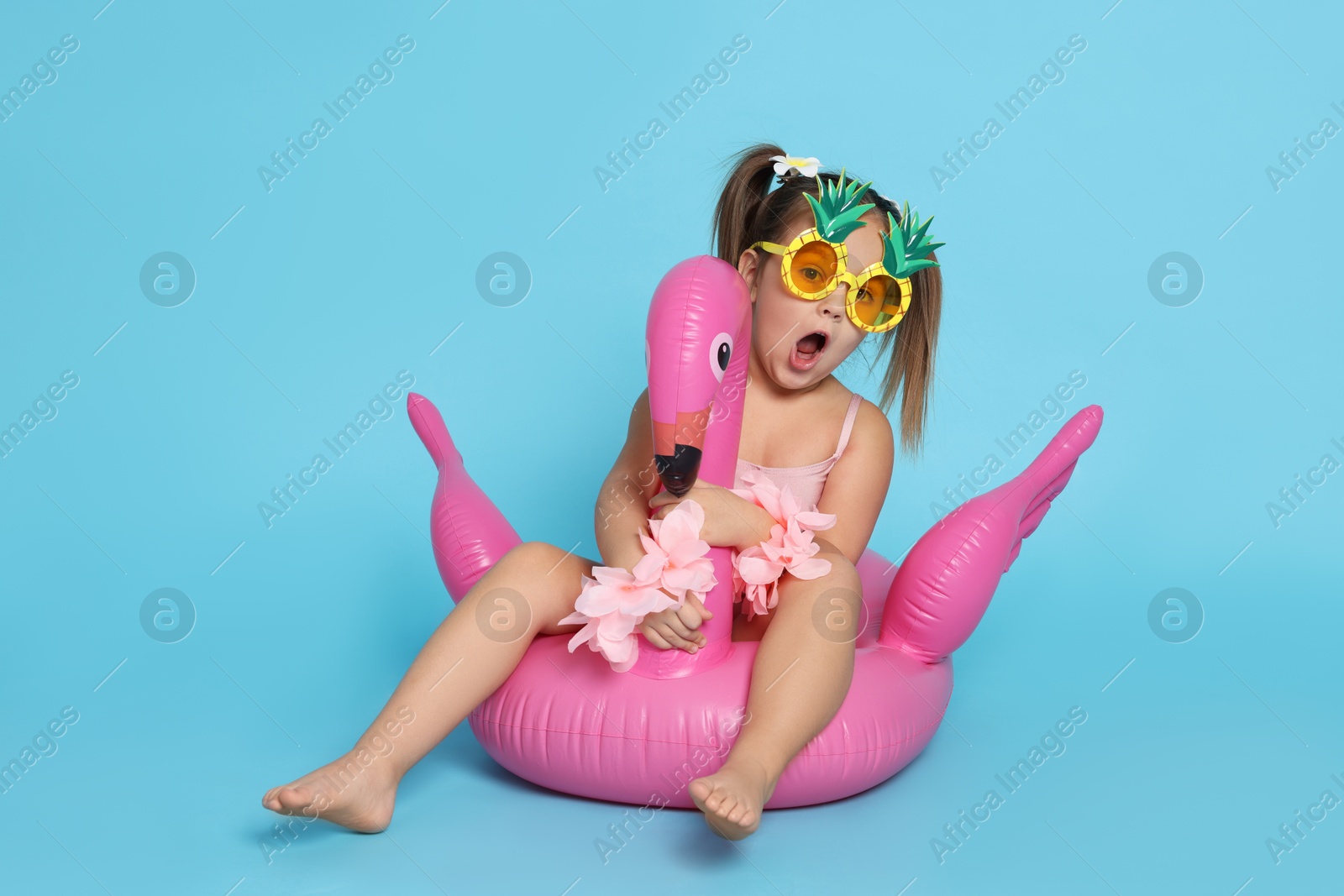 Photo of Cute little girl in swimsuit with funny sunglasses and inflatable ring on light blue background