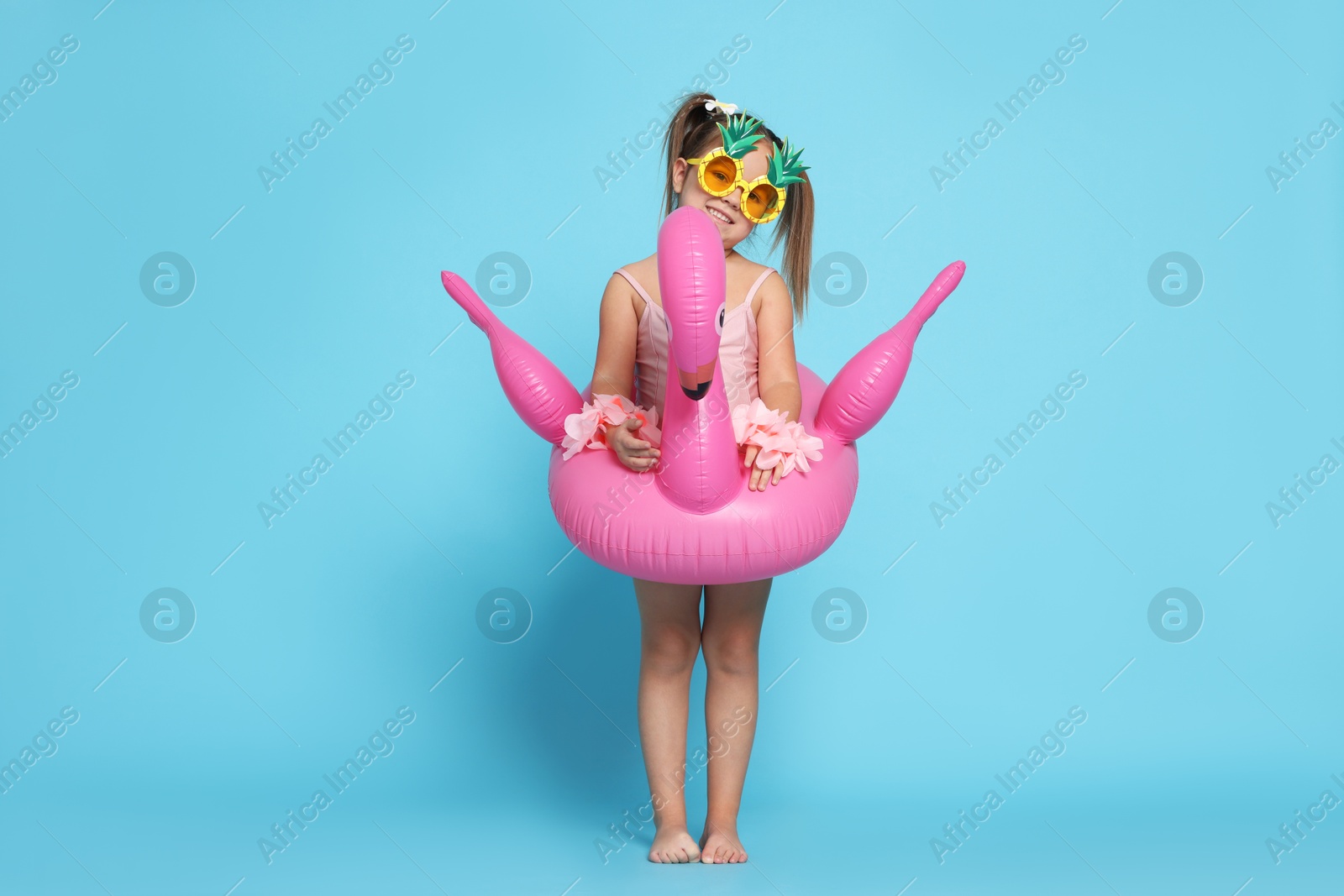 Photo of Cute little girl in swimsuit with funny sunglasses and inflatable ring on light blue background