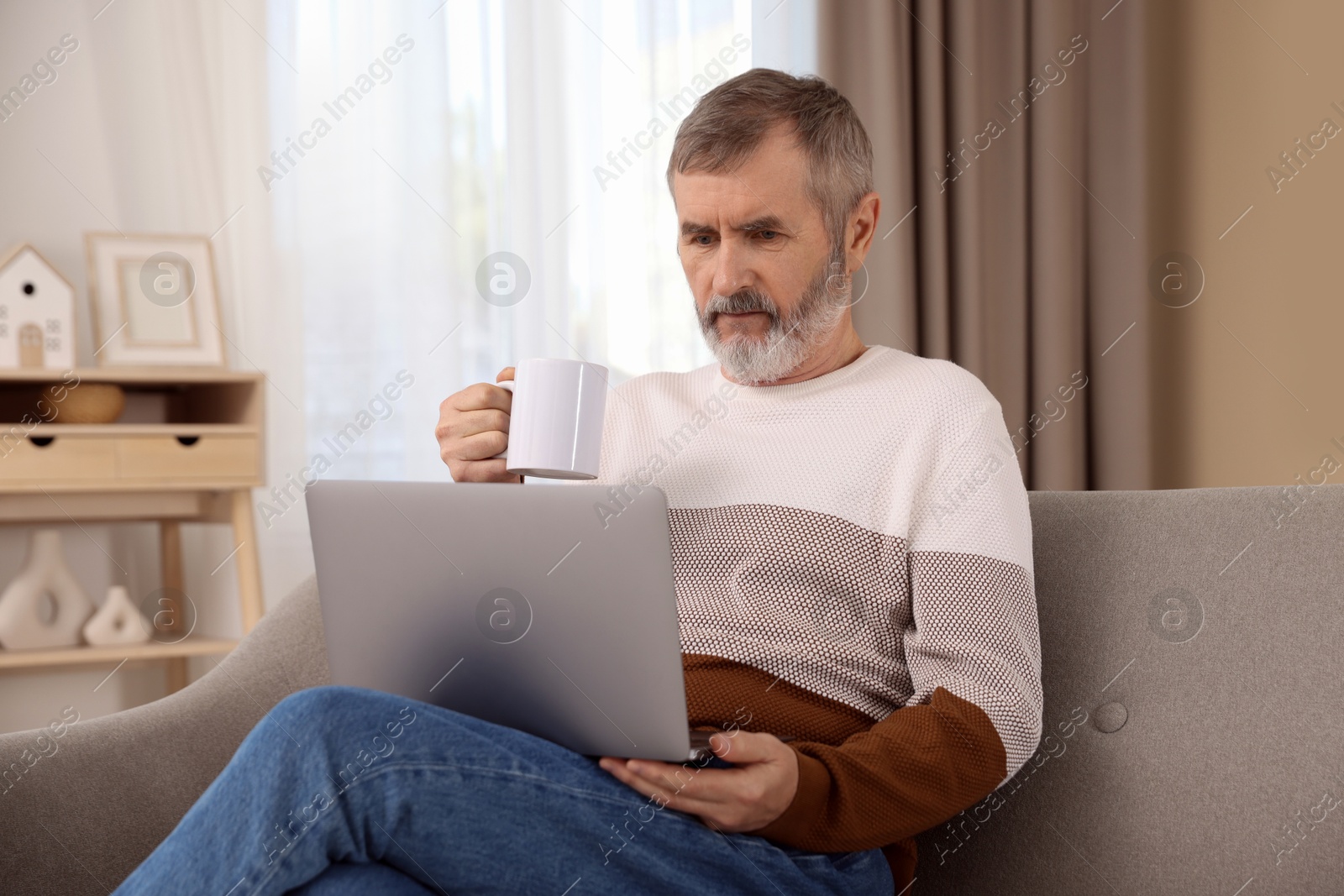 Photo of Mature man with laptop and cup of hot drink on sofa at home