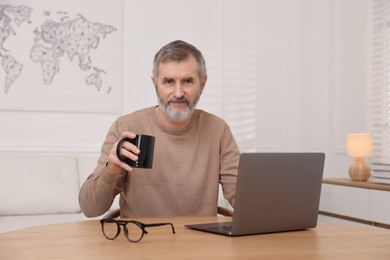 Photo of Mature man drinking coffee at table with laptop indoors