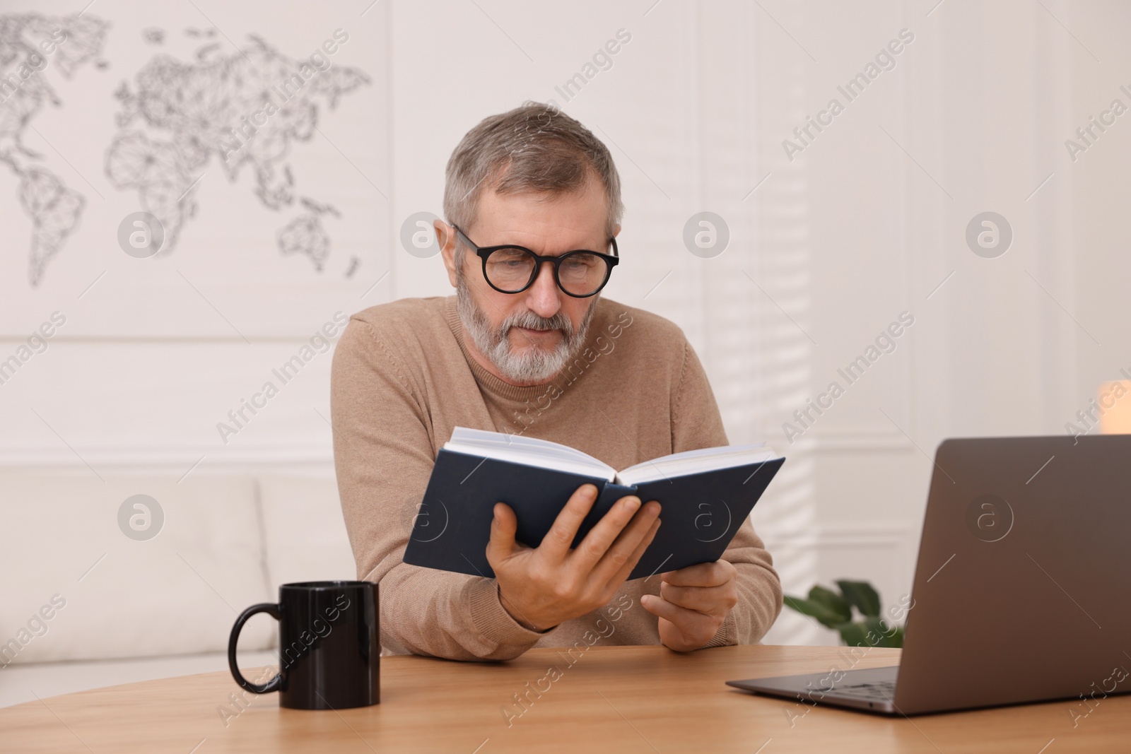 Photo of Mature man reading book at table with laptop indoors