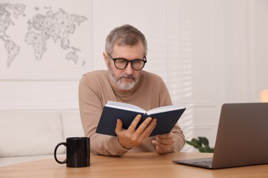 Mature man reading book at table with laptop indoors