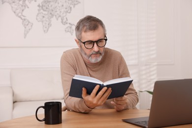 Mature man reading book at table with laptop indoors