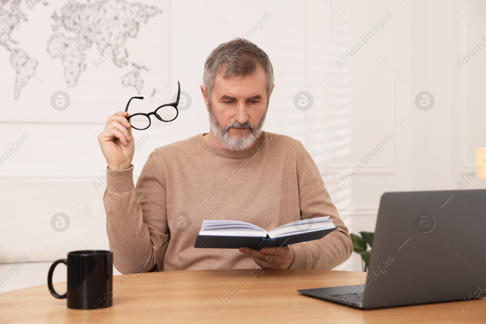 Photo of Mature man reading book at table with laptop indoors