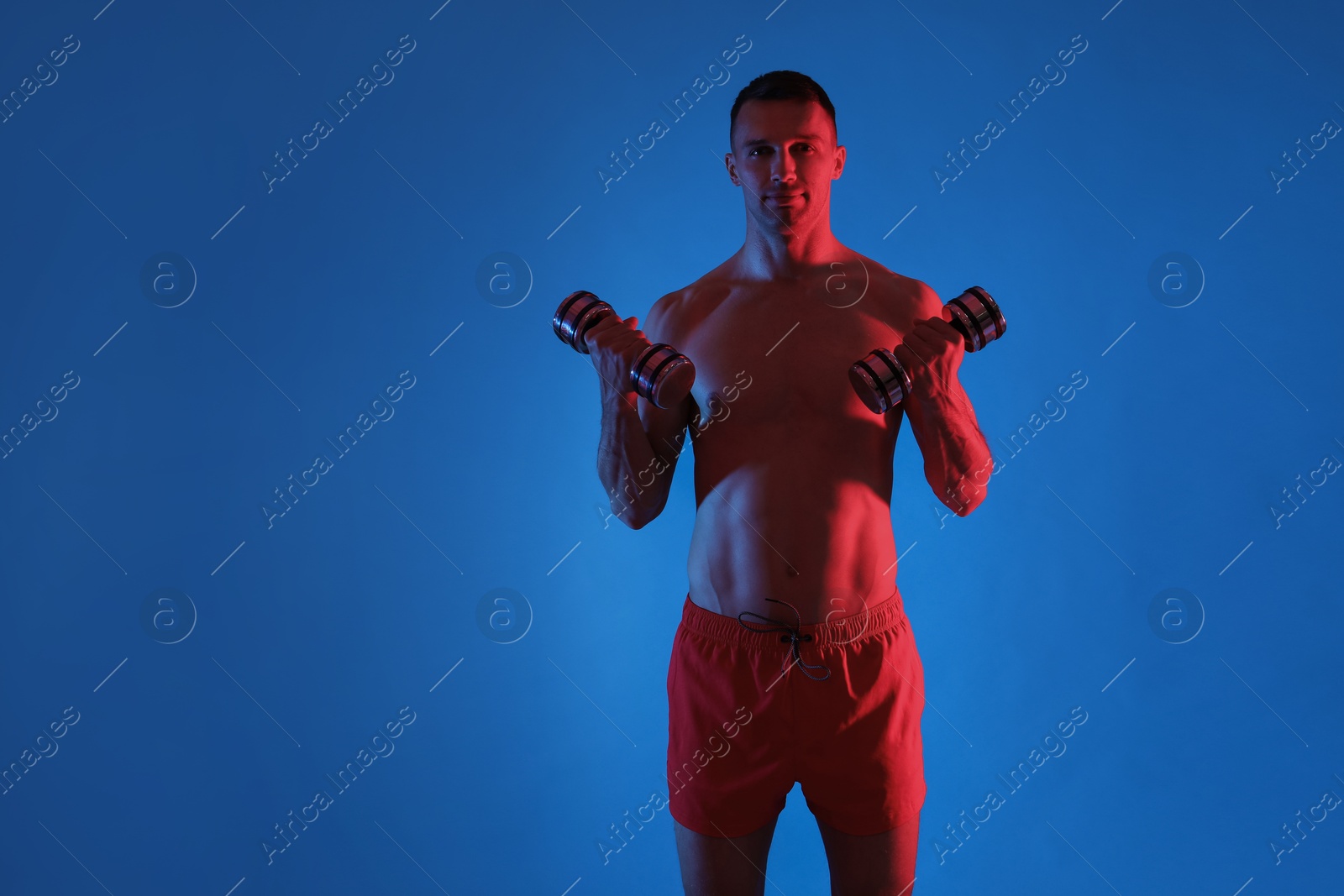 Photo of Man exercising with dumbbells in red light on blue background. Space for text