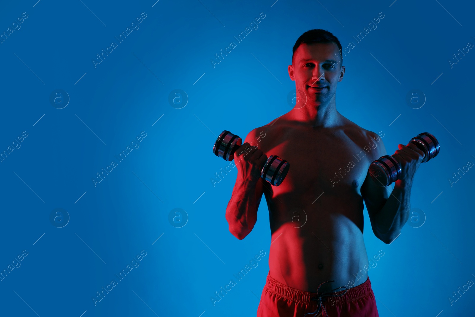 Photo of Man exercising with dumbbells in red light on blue background, space for text