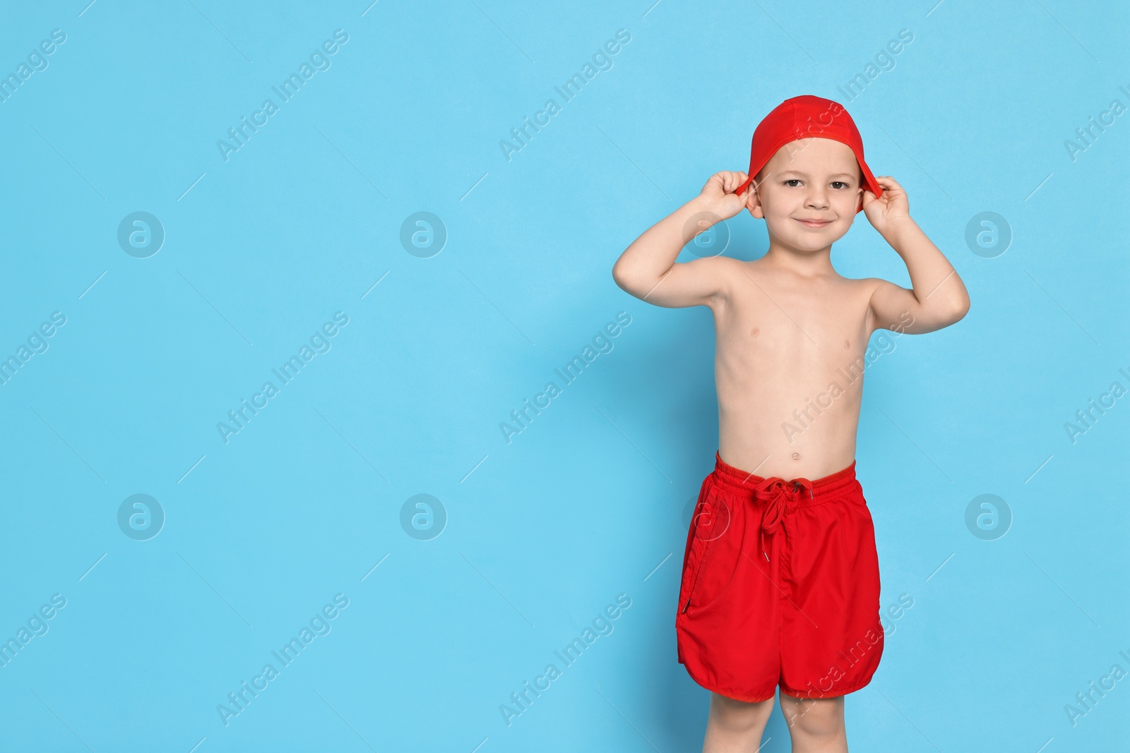 Photo of Cute little boy in beachwear with swim cap on light blue background. Space for text