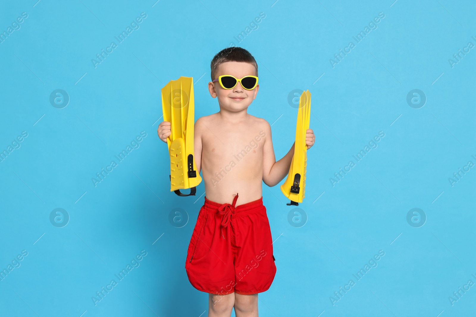 Photo of Cute little boy in beachwear with sunglasses and fins on light blue background