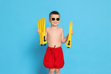 Cute little boy in beachwear with sunglasses and fins on light blue background