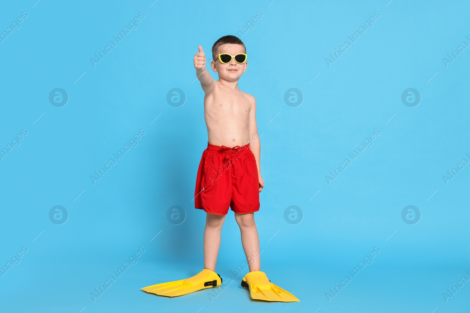 Photo of Cute little boy in beachwear with sunglasses and fins on light blue background