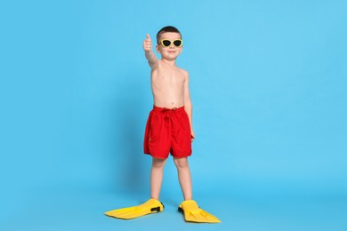 Photo of Cute little boy in beachwear with sunglasses and fins on light blue background