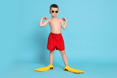 Photo of Cute little boy in beachwear with sunglasses and fins on light blue background