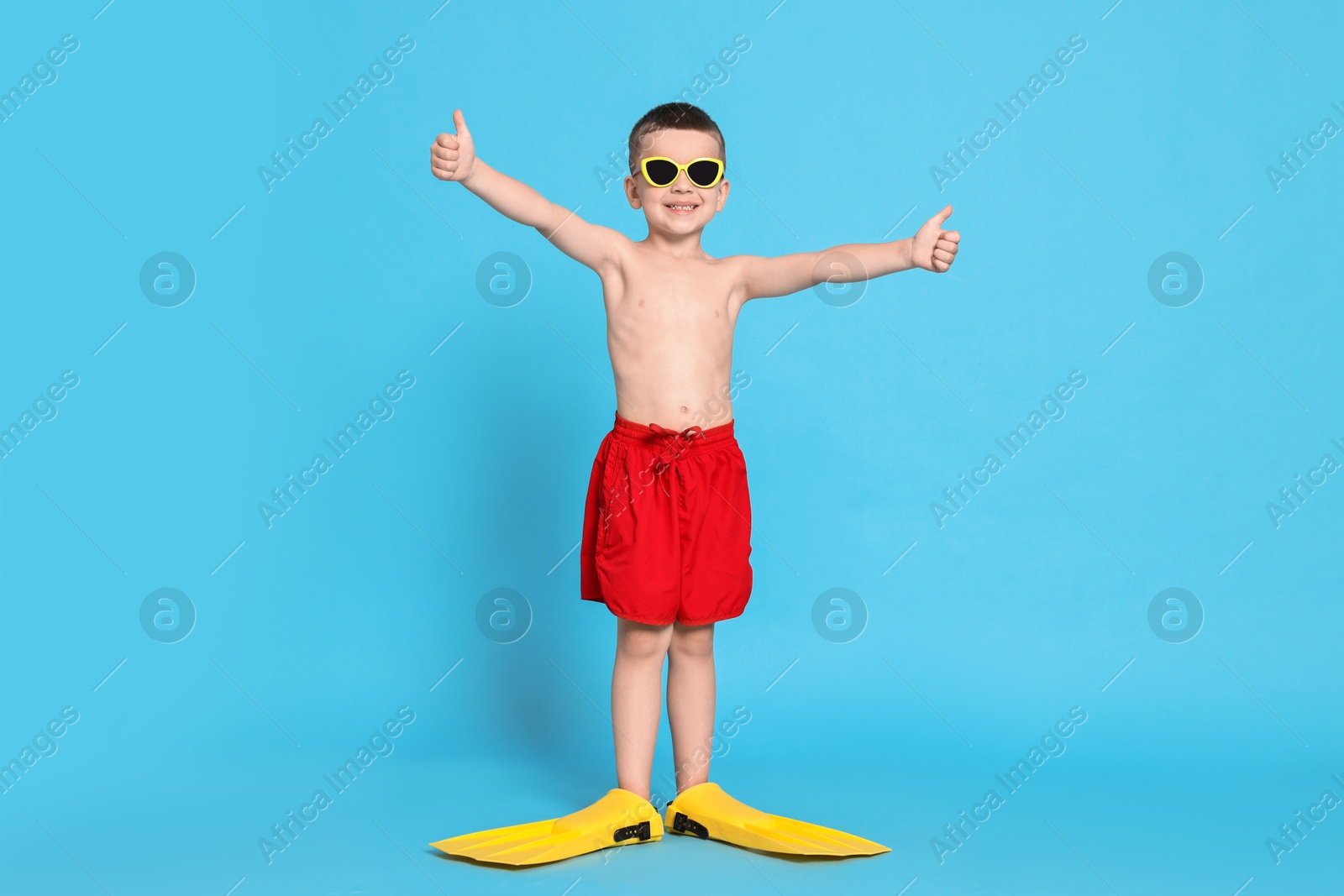 Photo of Cute little boy in beachwear with sunglasses and fins on light blue background