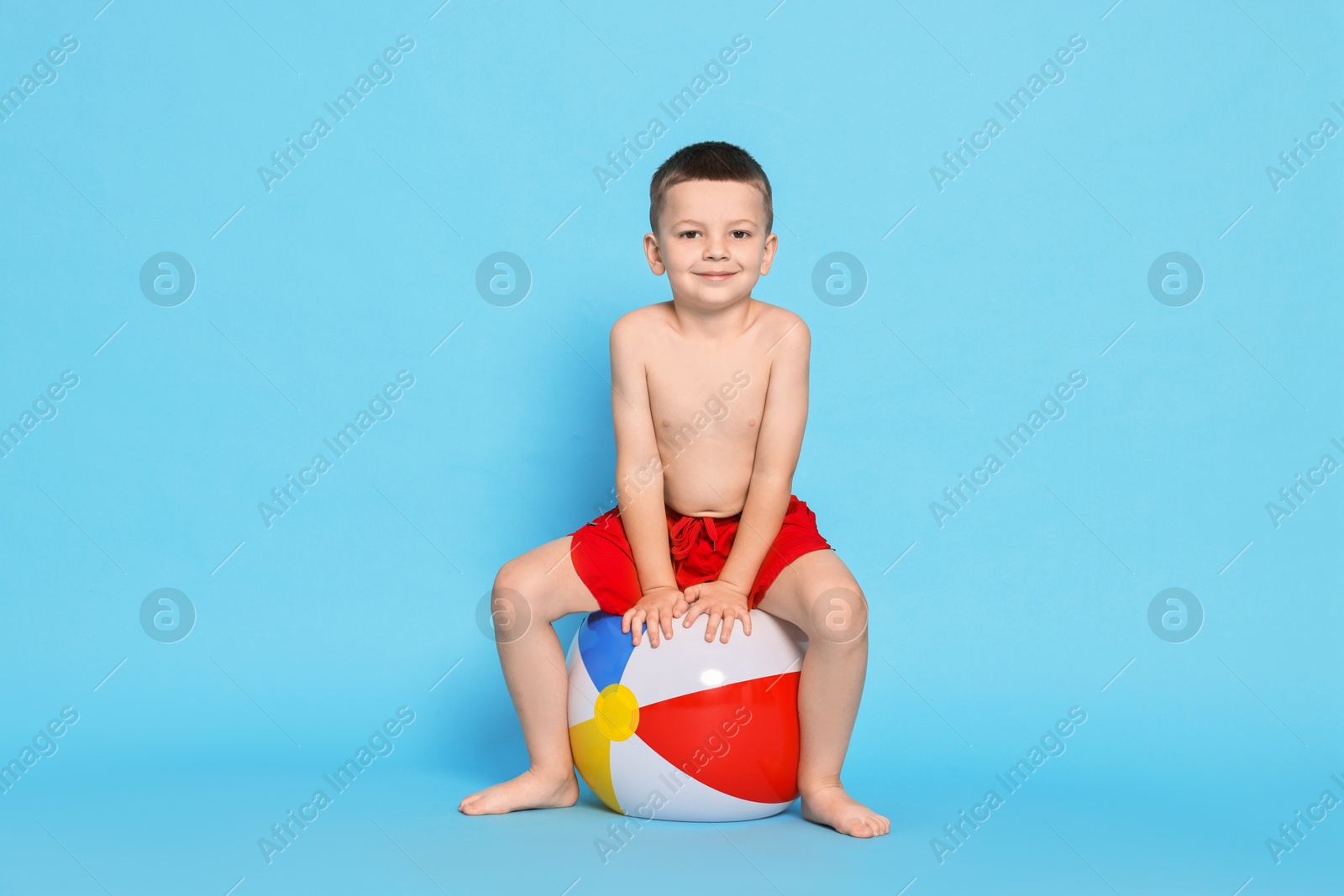 Photo of Cute little boy in beachwear with inflatable ball on light blue background