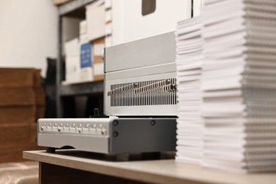 Photo of Modern binding machine and stacks of notebooks on wooden table indoors, selective focus