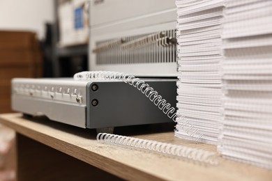 Photo of Modern binding machine, stacks of notebooks and double loop wire binding spines on wooden table indoors, closeup