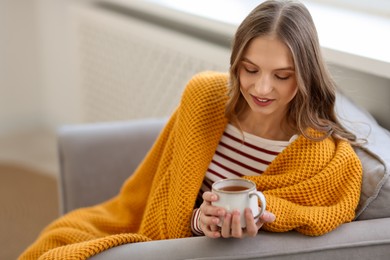 Charming young woman with cup of hot drink on sofa at home, space for text. Autumn atmosphere