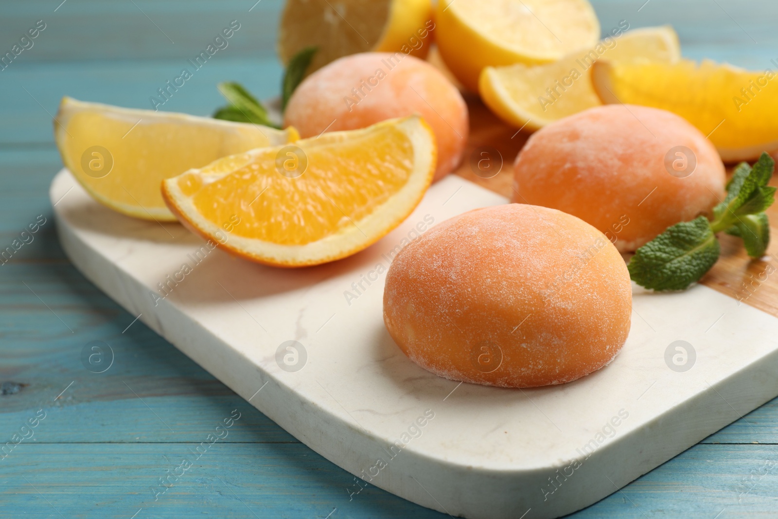 Photo of Board with tasty mochi, cut orange and mint on light blue wooden table, closeup