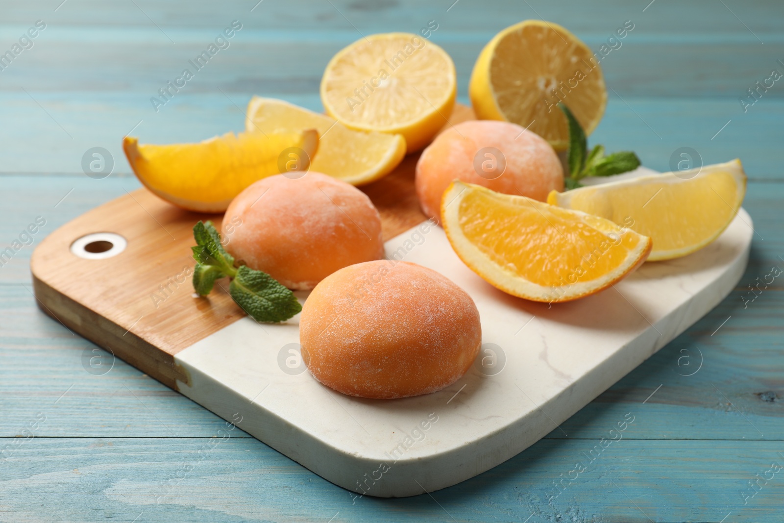 Photo of Board with tasty mochi, cut orange, lemon and mint on light blue wooden table, closeup