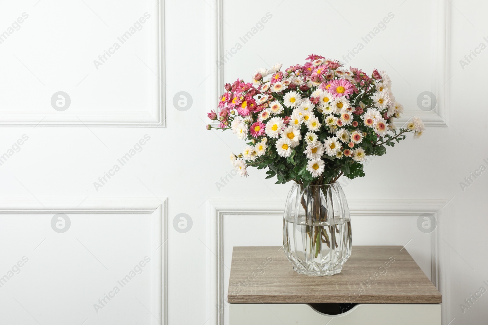 Photo of Vase with beautiful flowers on wooden nightstand near white wall, space for text