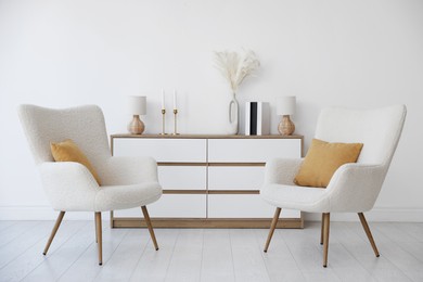 Soft armchairs and chest of drawers near white wall indoors