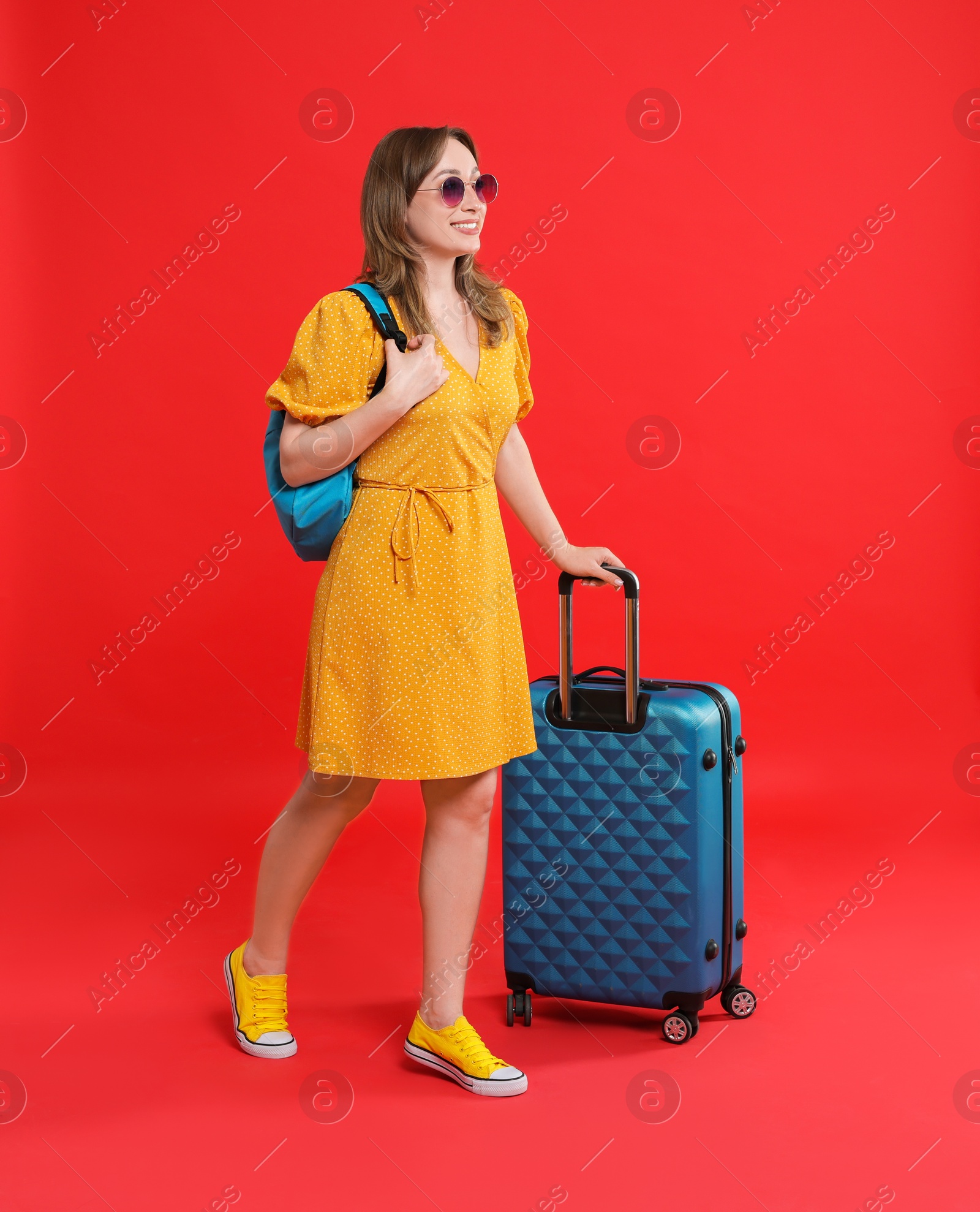 Photo of Happy traveller in sunglasses with suitcase on red background