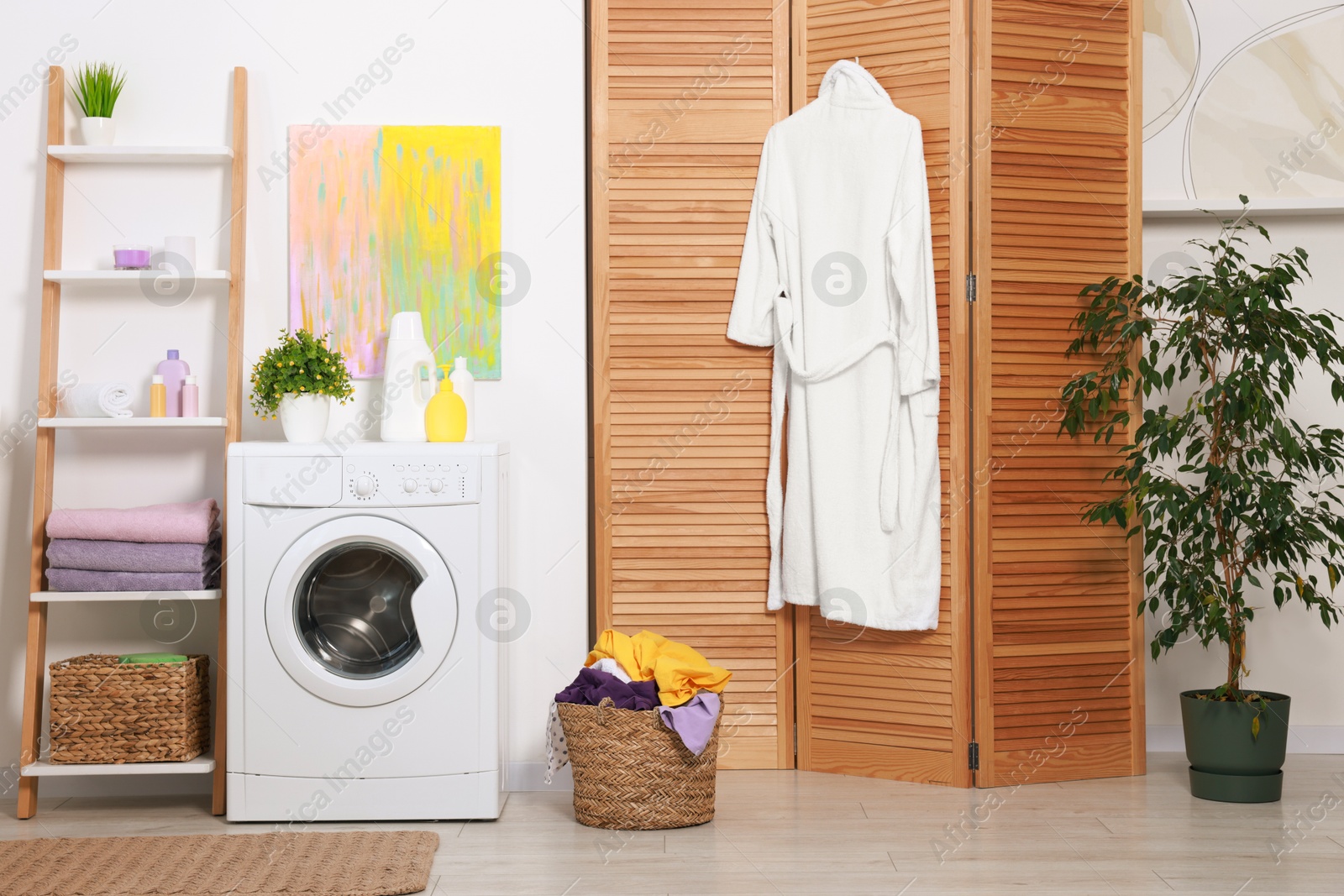 Photo of Washing machine, detergents, bathrobe, clean towels and basket with laundry in room