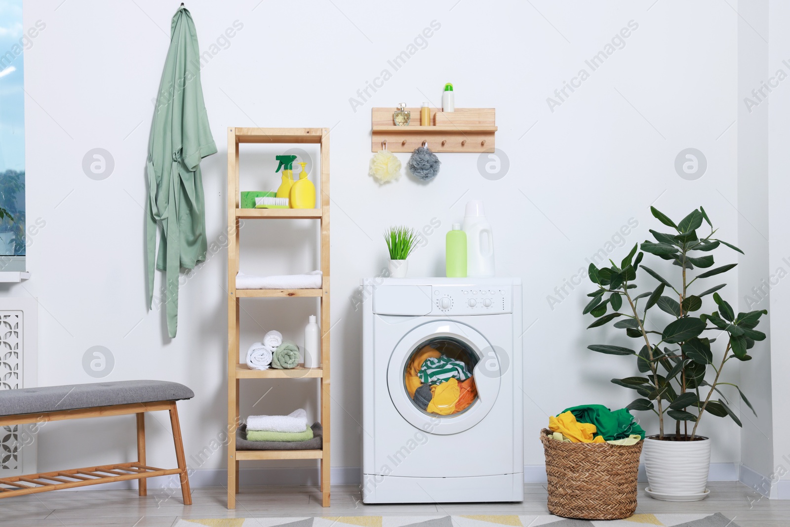 Photo of Washing machine, detergents, bathrobe and basket with laundry in room