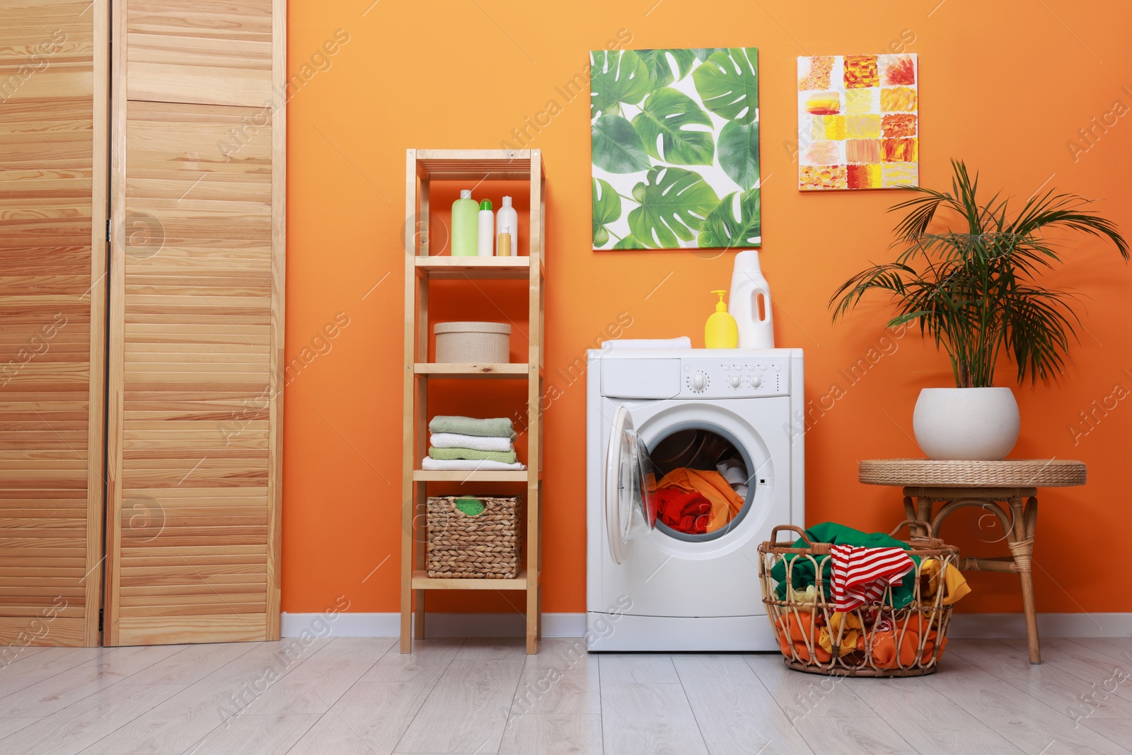 Photo of Washing machine, detergents, clean towels and basket with laundry near orange wall indoors