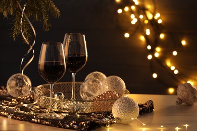 Photo of Glasses with red wine and beautiful Christmas decor on white table against blurred lights