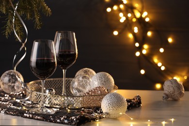 Photo of Glasses with red wine and beautiful Christmas decor on white table against blurred lights