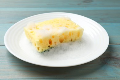 Photo of Yellow sponge with foam and plate on light blue wooden table, closeup