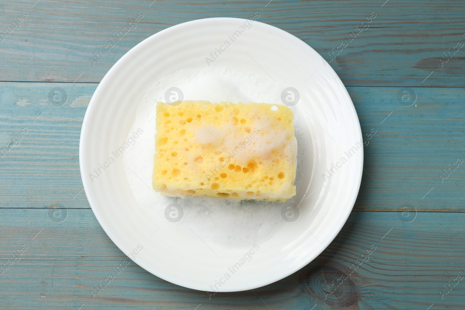 Photo of Yellow sponge with foam and plate on light blue wooden table, top view