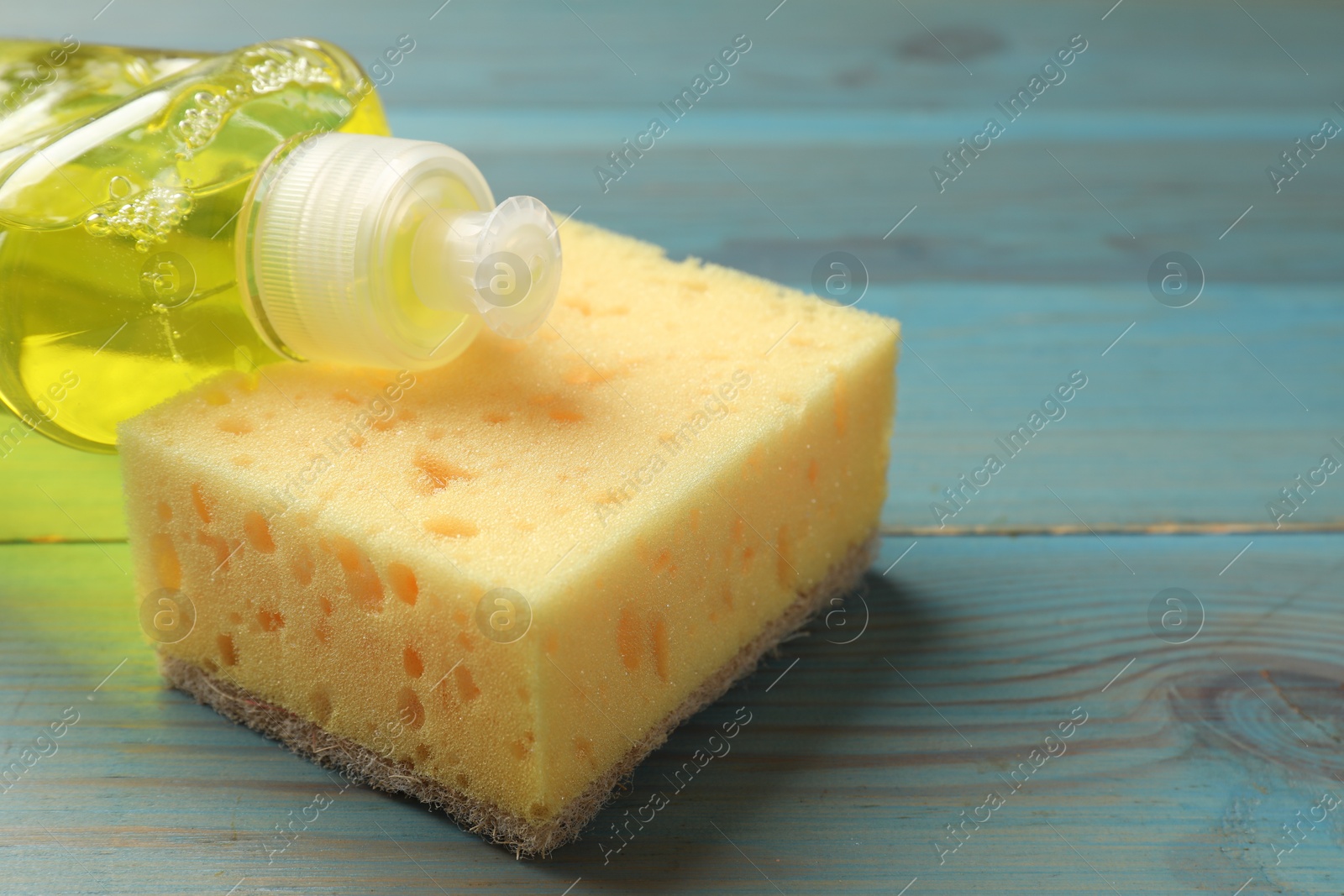 Photo of Yellow sponge and dish soap on light blue wooden table, closeup. Space for text