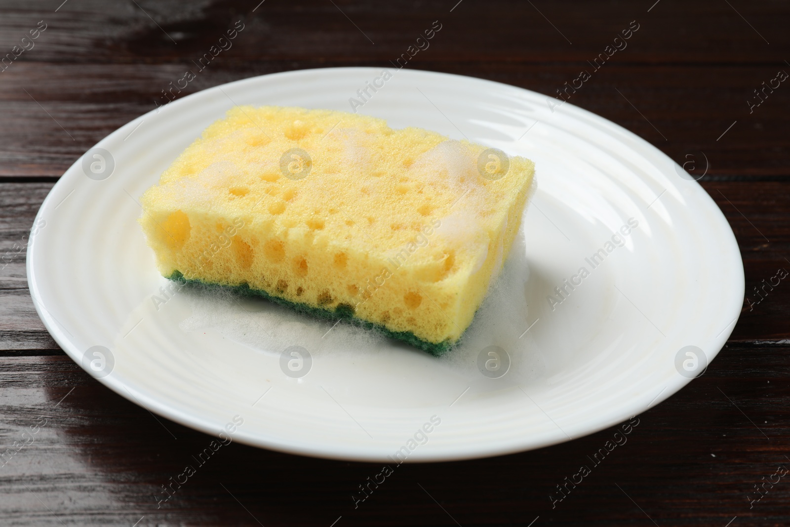 Photo of Yellow sponge with foam and plate on wooden table, closeup