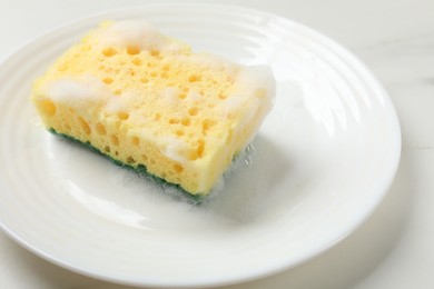 Photo of Yellow sponge with foam and plate on white table, closeup