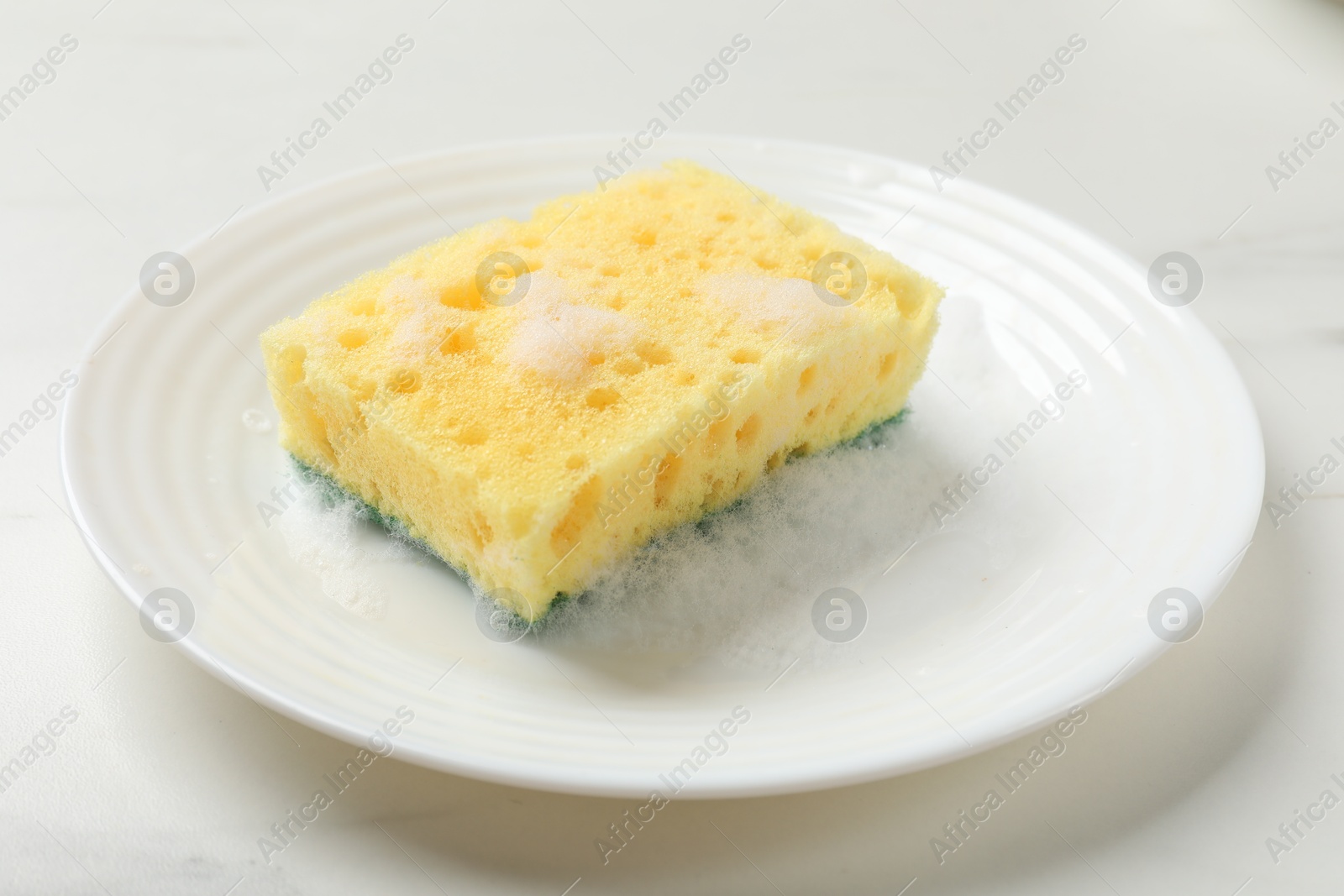 Photo of Yellow sponge with foam and plate on white table, closeup