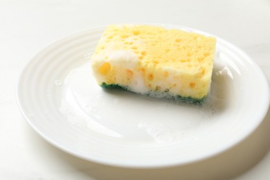 Photo of Yellow sponge with foam and plate on white table, closeup