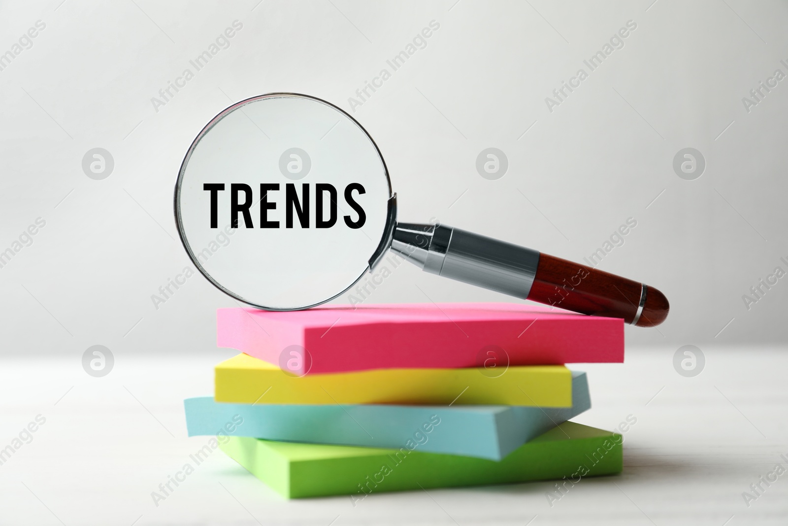 Image of Word Trends in magnifying glass on stack of paper notes against white background, closeup. Concept of searching for new and popular ideas