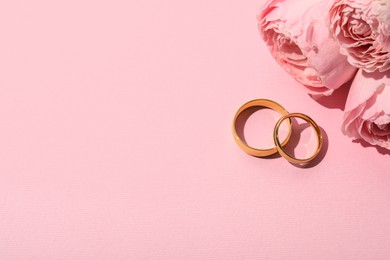 Photo of Golden wedding rings and flowers on pink background, space for text