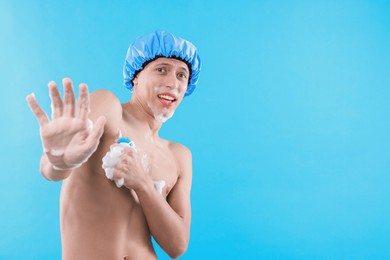 Photo of Emotional man with shower cap and mesh sponge making stop gesture on light blue background, space for text