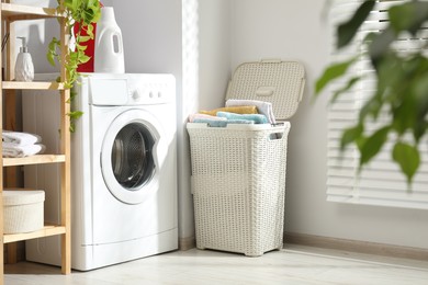 Photo of Wicker basket full of laundry, washing machine and detergents in bathroom