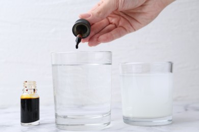 Photo of Iodine starch test. Woman dripping aqueous iodine into glass of water at white marble table, closeup