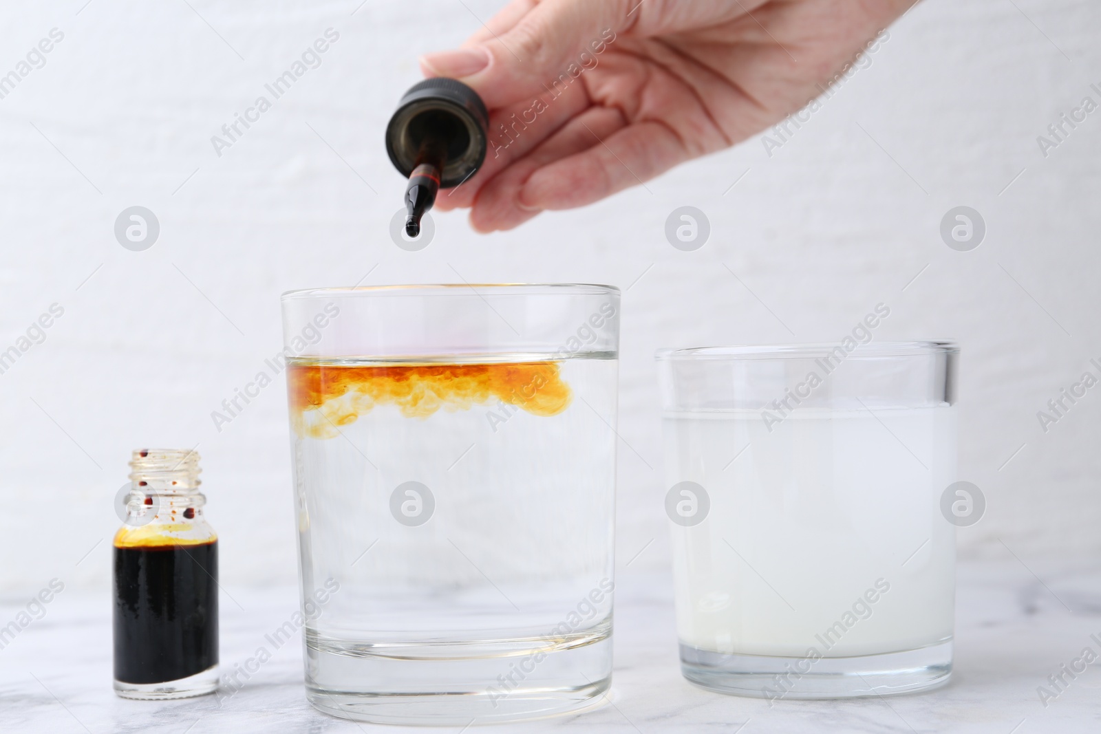 Photo of Iodine starch test. Woman dripping aqueous iodine into glass of water at white marble table, closeup