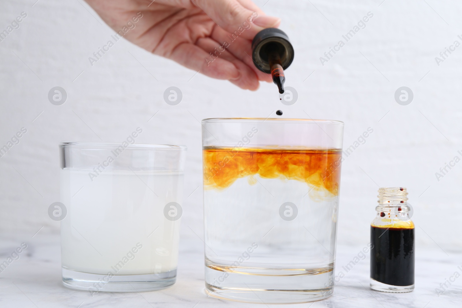 Photo of Iodine starch test. Woman dripping aqueous iodine into glass of water at white marble table, closeup