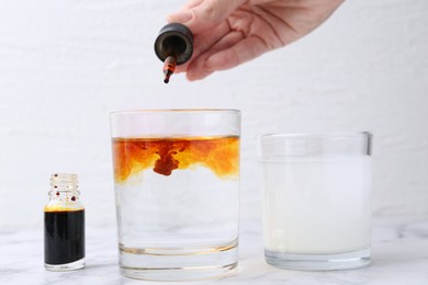 Photo of Iodine starch test. Woman dripping aqueous iodine into glass of water at white marble table, closeup