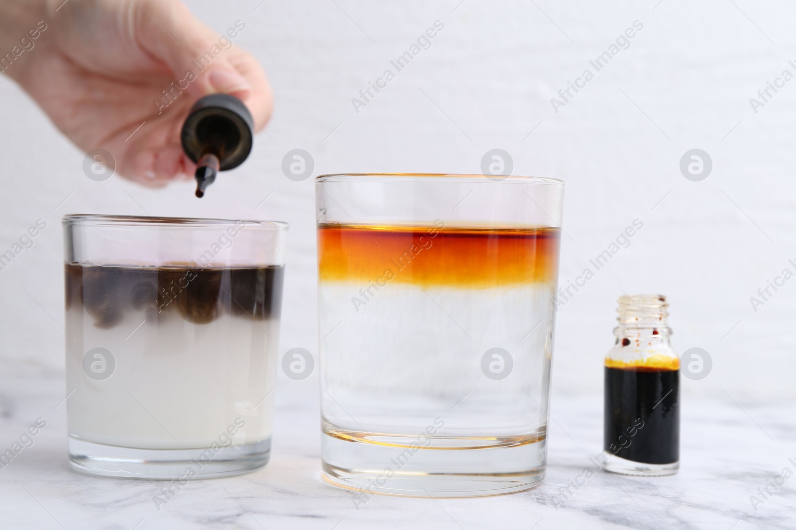 Photo of Iodine starch test. Woman dripping aqueous iodine into glass of water containing starch at white marble table, closeup