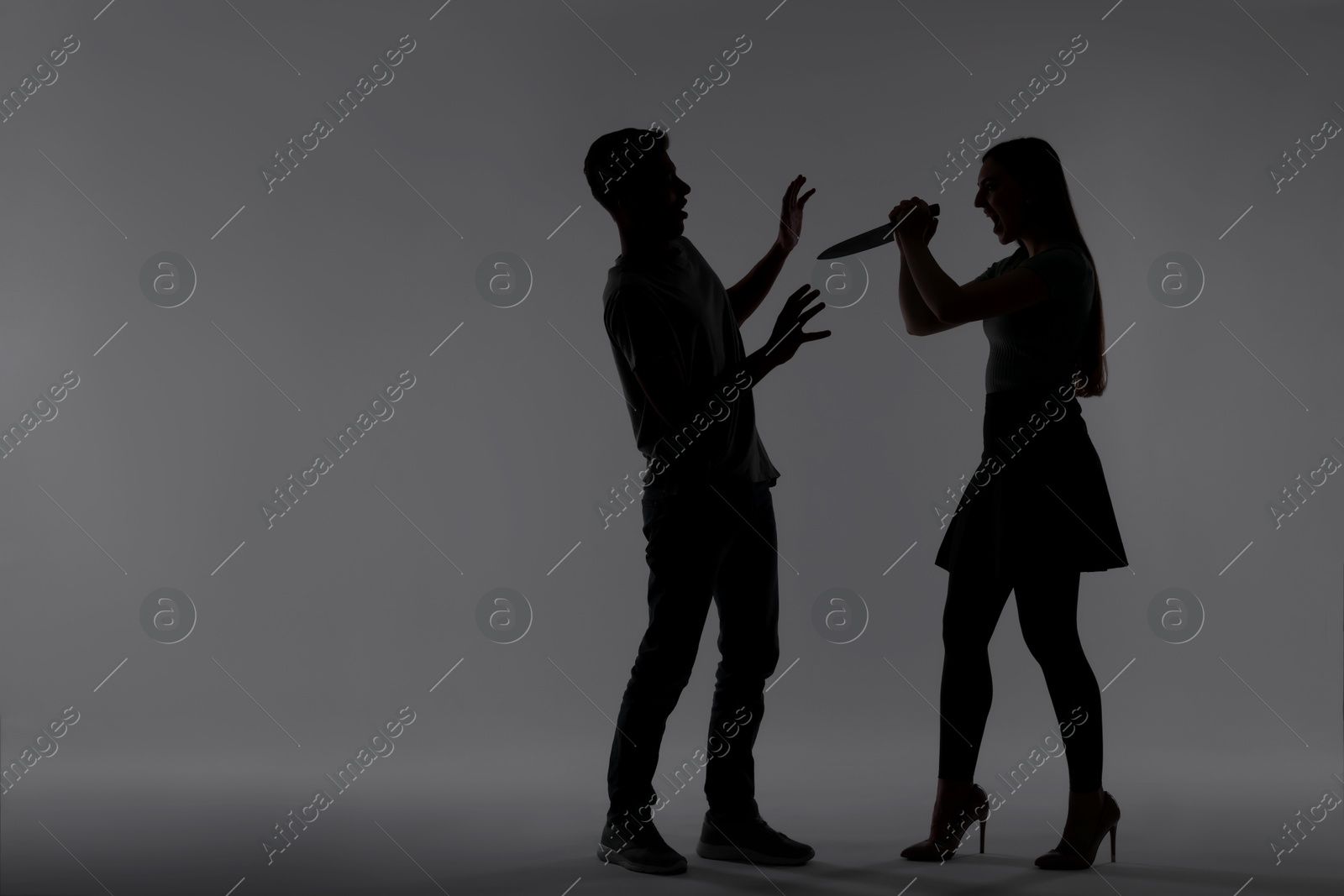 Photo of Domestic violence. Silhouette of woman with knife threatening her husband on grey background