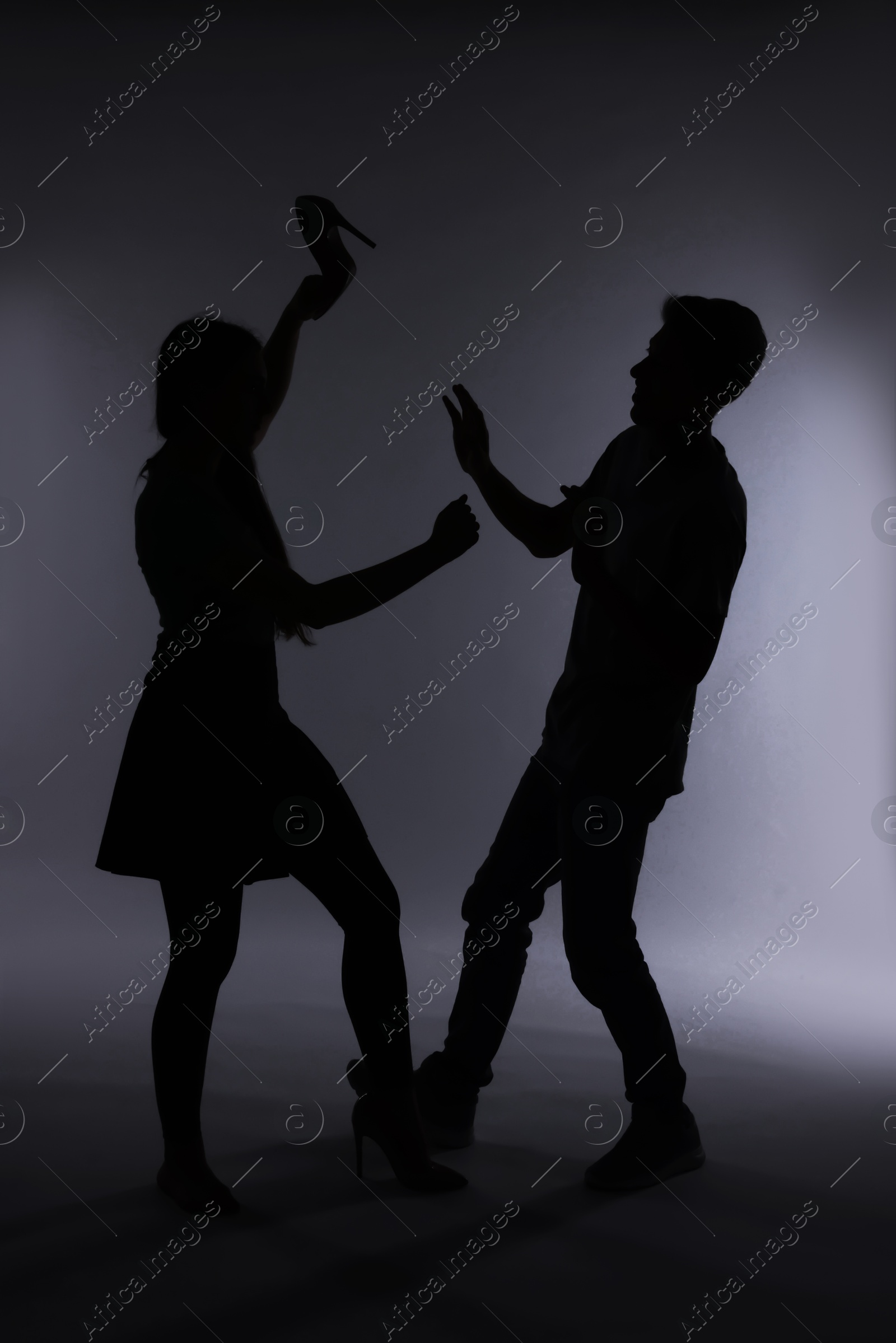 Photo of Domestic violence. Silhouette of woman with high-heeled shoe threatening her husband on grey background