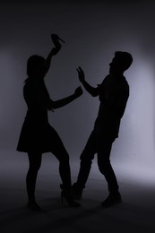 Domestic violence. Silhouette of woman with high-heeled shoe threatening her husband on grey background